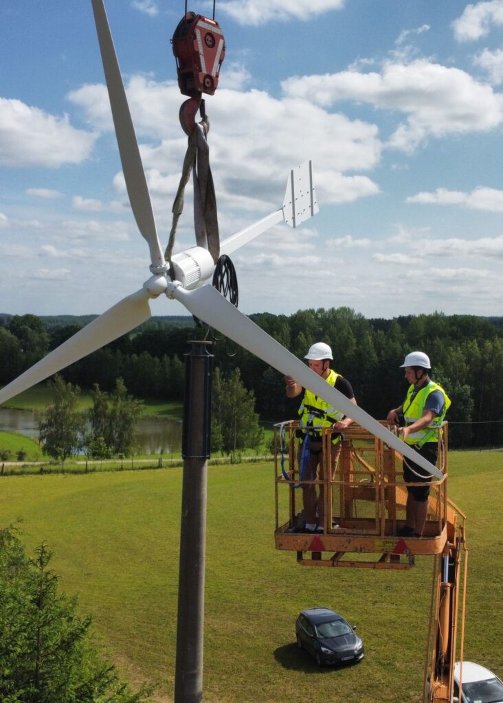 Dwóch mężczyzn współpracuje przy instalacji turbiny wiatrowej na otwartym polu, prezentując wysiłki w zakresie energii odnawialnej.