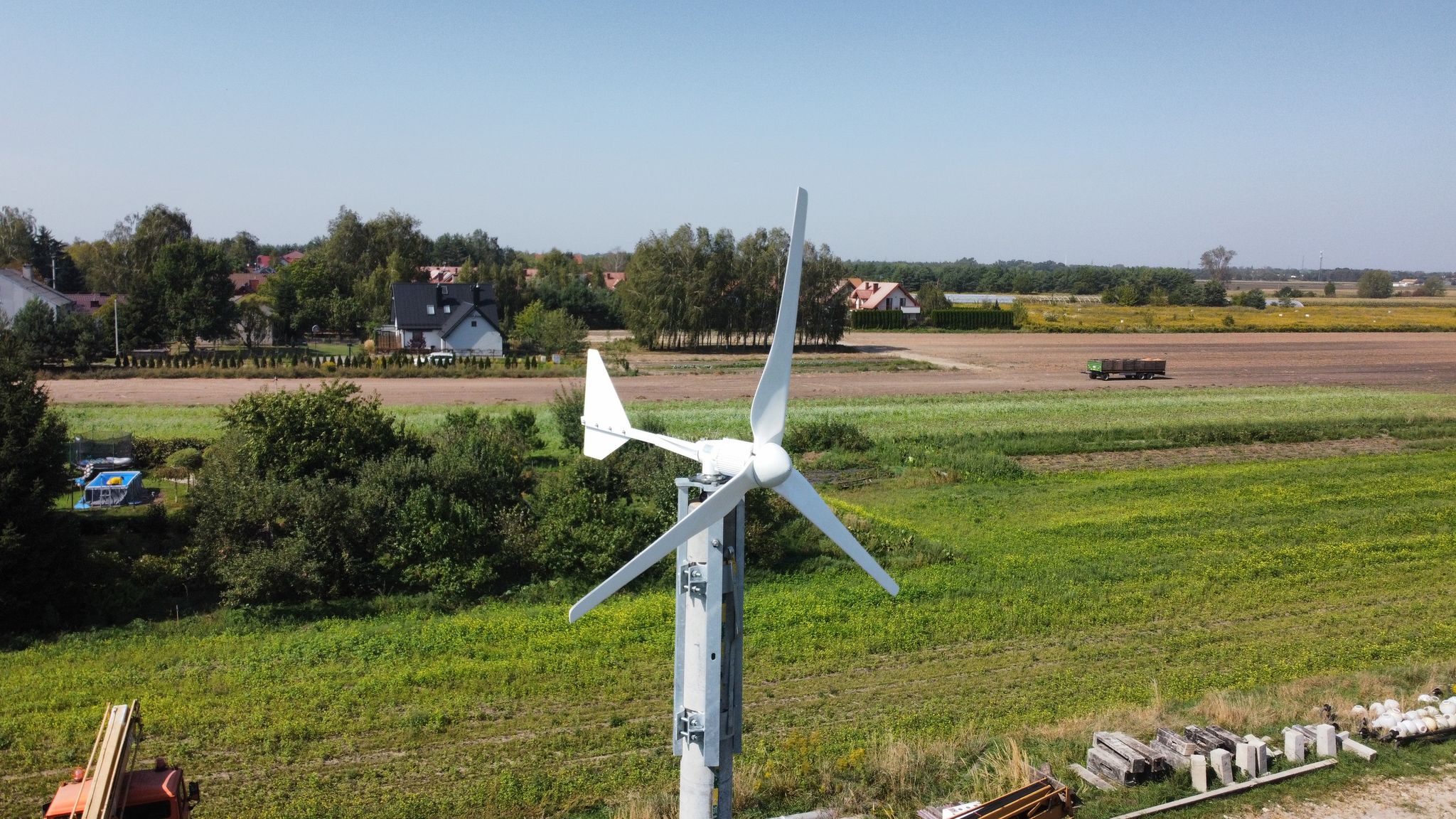 Turbina wiatrowa stoi wysoko na polu, z domem widocznym w tle pod czystym, błękitnym niebem.
